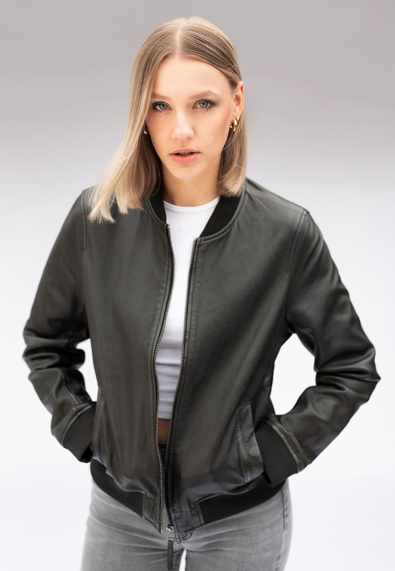 Frau mit glattem blondem Haar, die goldene Creolenohrringe, eine schwarze Lederjacke, ein weißes Hemd und graue Jeans trägt, steht mit Händen in den Taschen.