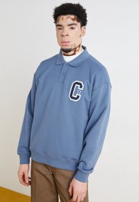 Polo shirt bleu à manches longues avec un col, comportant une lettre "C" brodée en blanc sur la poitrine gauche et des poignets côtelés. Porté avec un pantalon beige.