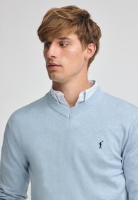 Ljusblå V-ringad tröja i mjukt stickat material. Ribbad krage och ärmslut, med en liten svart logotyp på vänster bröst.