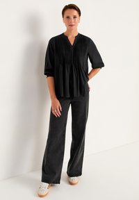 Schwarze, plissierte Bluse mit V-Ausschnitt und 3/4-Ärmeln, kombiniert mit dunkelgrauen, weiten Jeans und weißen Sneakers. Lässiger Schnitt.