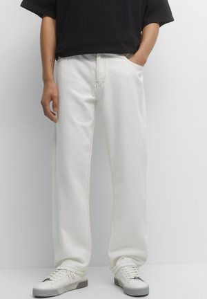 Personne portant un pantalon blanc ample et des baskets blanches, avec une main dans la poche et une chemise noire à manches courtes partiellement visible.