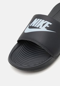 Μαύρη σανδάλι Nike με απαλό, υφασμένο λουράκι και λογότυπο σε γαλάζιο χρώμα. Η επιφάνεια για τα πόδια διαθέτει ραβδωτές λεπτομέρειες για καλύτερη πρόσφυση.