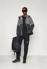 Oseba nosi sončna očala, sivo-črno jakno, črne hlače in škornje ter drži velik črn nahrbtnik z znamko The North Face.