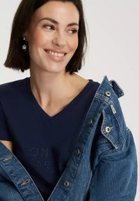 Granatowa koszulka z dekoltem w kształcie litery V z nadrukiem w kropki, założona pod jasnoniebieską kurtką dżinsową z srebrnymi guzikami i miękką teksturą.