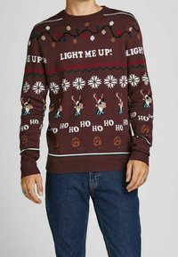Pull marron avec des motifs festifs, incluant des rennes, des flocons de neige et des biscuits en pain d'épice. Présente le texte "LIGHT ME UP!" en crème.