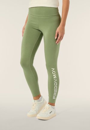 Leggings verdi con una vita alta e una texture liscia. Marchio bianco ben visibile sulla gamba inferiore. Abbinate a scarpe sportive bianche.