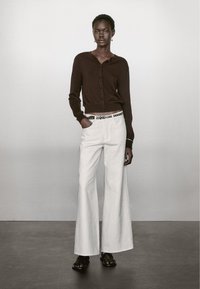 Cardigan corto marrone con bottoni abbinato a pantaloni bianchi a gamba larga con una cintura in contrasto. Calzature: sandali neri. Design semplice e moderno.