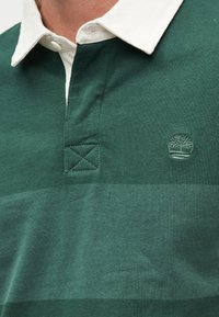 Σκούρο πράσινο ράγκμπι πουκάμισο με φαρδιές ρίγες, λευκό γιακά, κουμπωτή λωρίδα κουμπιών και ένα μικρό κεντημένο λογότυπο δέντρου στο στήθος.