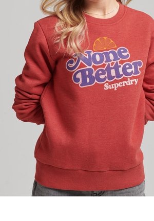 Kobieta w rdzawobrązowej bluzie z napisem "None Better Superdry" i grafiką plastra pomarańczy na piersi.