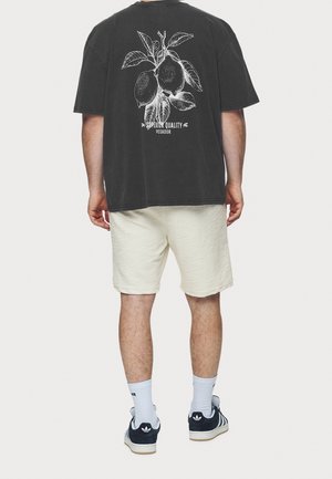 Osoba nosząca czarną oversize koszulkę z białą ilustracją gałązki cytryny oraz tekstem, jasne teksturowane szorty, białe skarpetki i granatowe sneakersy.
