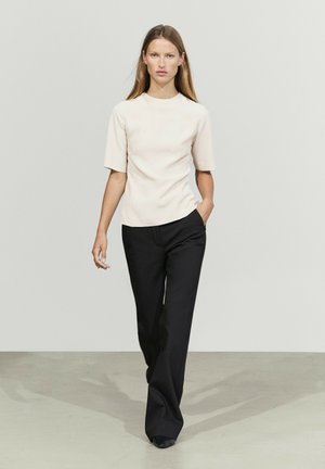 Femme marchant vers l'avant portant un haut beige à manches courtes et un pantalon noir à jambes larges sur un fond clair uni.
