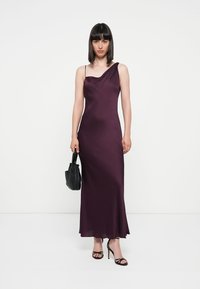 Paarse, mouwloze midi-jurk met een asymmetrisch ontwerp. Gladde, licht glanzende stof. Model houdt een kleine zwarte tas vast en draagt zwarte hakken.