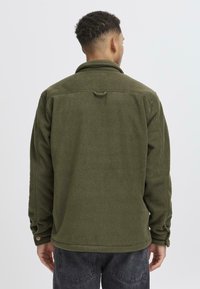 Olivengrüne Fleecejacke mit Kragen, geknöpften Ärmeln und einer Schlaufe hinten. Strukturierter Stoff, lockerer Schnitt.