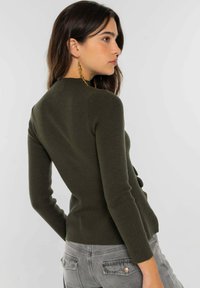 Pull ajusté vert olive avec manches longues et col montant. Présente un tricot texturé et un détail de boutons dans le dos.