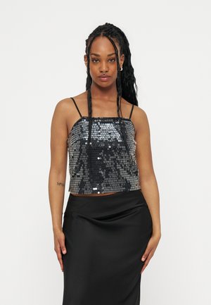 Νέα γυναίκα με μακριά πλεγμένα μαλλιά, φοράει μαύρο τοπ με λεπτές τιράντες διακοσμημένο με παγέτες και μαύρη φούστα, στέκεται μπροστά σε ένα απλό ανοιχτό φόντο.