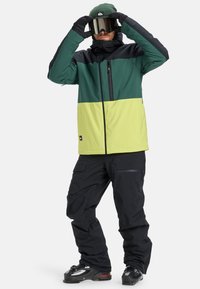 Men's snow jacket in green and yellow with black accents, featuring a zippered pocket. Paired with black snow pants and ski goggles.