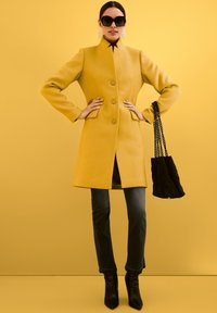 Geel wollen jas met een staande kraag en knopen aan de voorkant, gecombineerd met grijze jeans en zwarte puntige laarzen. Zwarte handtas met kettinghengsels.