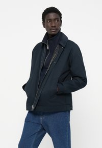 Navy blue jacket with a zip closure, snap buttons, side pockets, and a contrasting collar. Lined with a green plaid pattern.