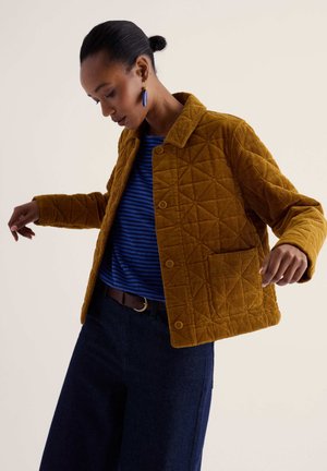 Femme portant une veste matelassée en velours côtelé moutarde par-dessus un haut rayé bleu, pantalon foncé, avec des boucles d'oreilles bleues pendantes et les cheveux en chignon, posant avec les bras ouverts.