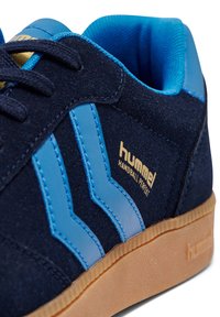 Navy suede sneaker featuring light blue leather accents and three stripes design. Gold branding label "hummel Handball Perfect" on side. Rubber sole.