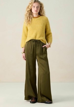 Vrouw staat met een gele donzige trui, olijfgroene wijde broek, schoenen met luipaardprint, amberkleurige ketting en gouden armbanden tegen een effen achtergrond.