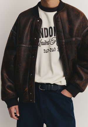 Rjava bomber jakna iz umetnega usnja z rebrastim ovratnikom in manšetami, nošena preko krem majice s črnim napisom, kombinirana s temno modrimi hlačami in črnim pasom.