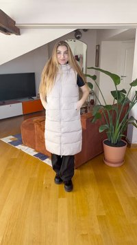 Polsterēts bēšs garš veste ar augstu apkakli, priekšā rāvējslēdzējs un kabatas, valkāta virs melnas blūzes, saskaņota ar melnām biksēm un apaviem.