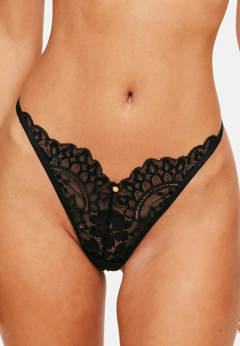 String en dentelle noire avec des motifs floraux, un bord festonné et un petit accent doré au centre avant. Bretelles latérales réglables.