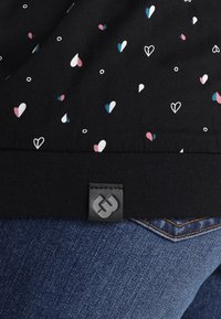 Czarna tkanina z małymi białymi, różowymi i niebieskimi wzorami serc i kółek, ryfrowanym wykończeniem oraz czarną metką powyżej niebieskich dżinsów.