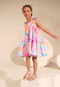 Vestido colorido de capas con tonos pastel de rosa, azul y naranja; presenta lazos en los hombros y un escote fruncido. Llevado con sandalias rosas.