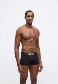 Fekete bokszerrövidnadrág vastag elasztikus derékpánttal, amelyen logó látható. Simább anyagfelület és testhezálló kialakítás emelik ki a test formáját.