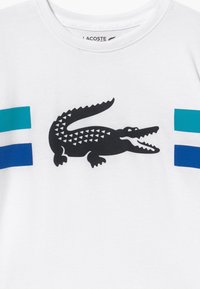 Bílý bavlněný T-shirt s černým logem krokodýla s trojúhelníkovými vzory a dvěma vodorovnými pruhy v tealové a modré barvě na rukávech.
