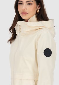 Cremefarbene wasserdichte Jacke mit abnehmbarer Kapuze, Frontreißverschluss und schwarzem runder Logo-Patch auf dem linken Ärmel. Glatte Textur.