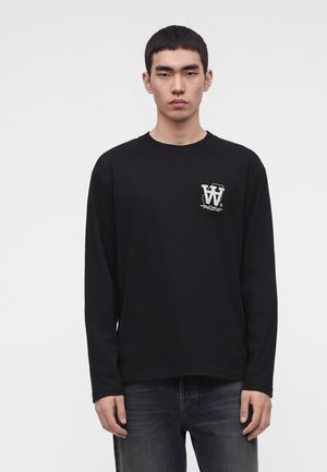 Jeune homme portant un t-shirt noir à manches longues avec un logo blanc sur la poitrine gauche et un jean gris foncé, debout devant un fond uni.