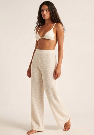 Mujer con cabello largo y rizado, vestida con un top de bikini de punto texturizado blanco y pantalones anchos a juego, de pie descalza sobre un fondo claro.