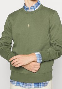 Muž v olivově zelené mikině přes modrou kostkovanou košili, se založenýma rukama, s béžovými kalhotami viditelnými v pase.