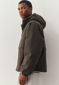 Olijfgroene jas met capuchon, elastische manchetten en een voorzak. Gladde stof met een matte afwerking, gedragen over grijze joggingbroek.