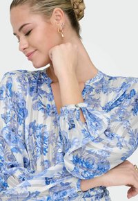 Femme aux cheveux blonds tressés, portant un chemisier bleu à fleurs et des boucles d'oreilles dorées, souriant les yeux fermés et la main près du menton.