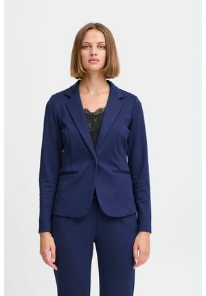 Femme aux cheveux lisses châtain clair portant un blazer bleu marine et un pantalon assorti sur un haut en dentelle noire, debout devant un fond blanc uni.