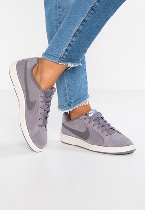 Stopy w szarych zamszowych sneakersach Nike z białymi podeszwami i niebieskich dżinsach z postrzępionym wykończeniem na białym tle.