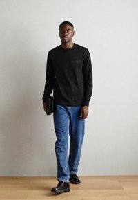 Černá košile s dlouhým rukávem s texturovanou látkou a malým logem. Nosit s modrými džíny a černými koženými botami, držící černý doplněk.