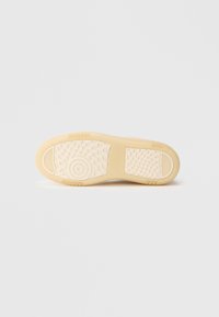 Podeszwa beżowego trampka z teksturowanymi wzorami chwytu, w tym okrągłymi i w kratkę, na jednolitym białym tle.