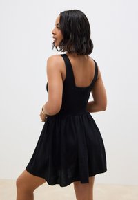 Zwarte mouwloze jurk met een aansluitend lijfje, taille die is ingesnoerd en een uitlopende rok. Gladde textuur, lengte tot halverwege de bovenbenen en een open rugontwerp.