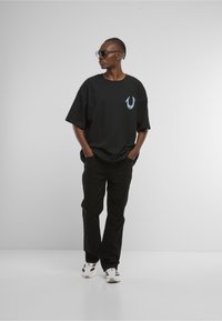 Czarny oversize t-shirt z białym i niebieskim logo na piersi, zestawiony z czarnymi spodniami i białymi sneakersami. Gładka tkanina i luźny krój.