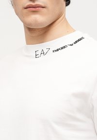 Κοντινό πλάνο ενός ατόμου που φοράει λευκό πουκάμισο με μαύρο κείμενο "EA7 Emporio Armani" τυπωμένο κοντά στον γιακά στη δεξιά πλευρά.