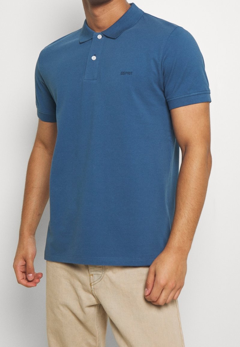 Homme portant un polo bleu à manches courtes avec deux boutons blancs et un pantalon beige devant un fond uni.
