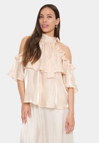 Blouson top in pale peach satin with ruffled layers, cold-shoulder design, and high neckline. Long, flowing sleeves.