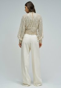 Eine Frau, die von hinten steht und ein goldenes, langärmliges, mit Pailletten besetztes Oberteil mit geraffter Taille sowie weite weiße Hosen trägt, vor einem einfarbigen grauen Hintergrund.