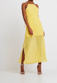 Robe maxi jaune avec un corsage ajusté, des bretelles spaghetti réglables et des fentes latérales. Confectionnée dans un tissu léger et fluide avec une superposition transparente.
