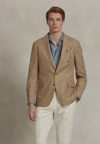 Jonge man in een beige geruite blazer, blauwe overhemd, witte broek, met een pochette en horloge, staand tegen een vlakke achtergrond.